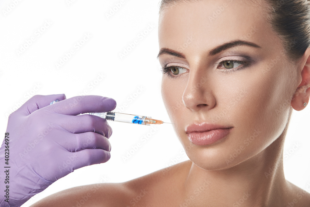 Beautician's hand in purple glove holding a syringe near beautiful woman's face. Portrait isolated on a white background. Anti-aging treatment. Beautician's hand in purple glove holding a syringe near beautiful woman's face. Portrait isolated on a white background. Anti-aging treatment.