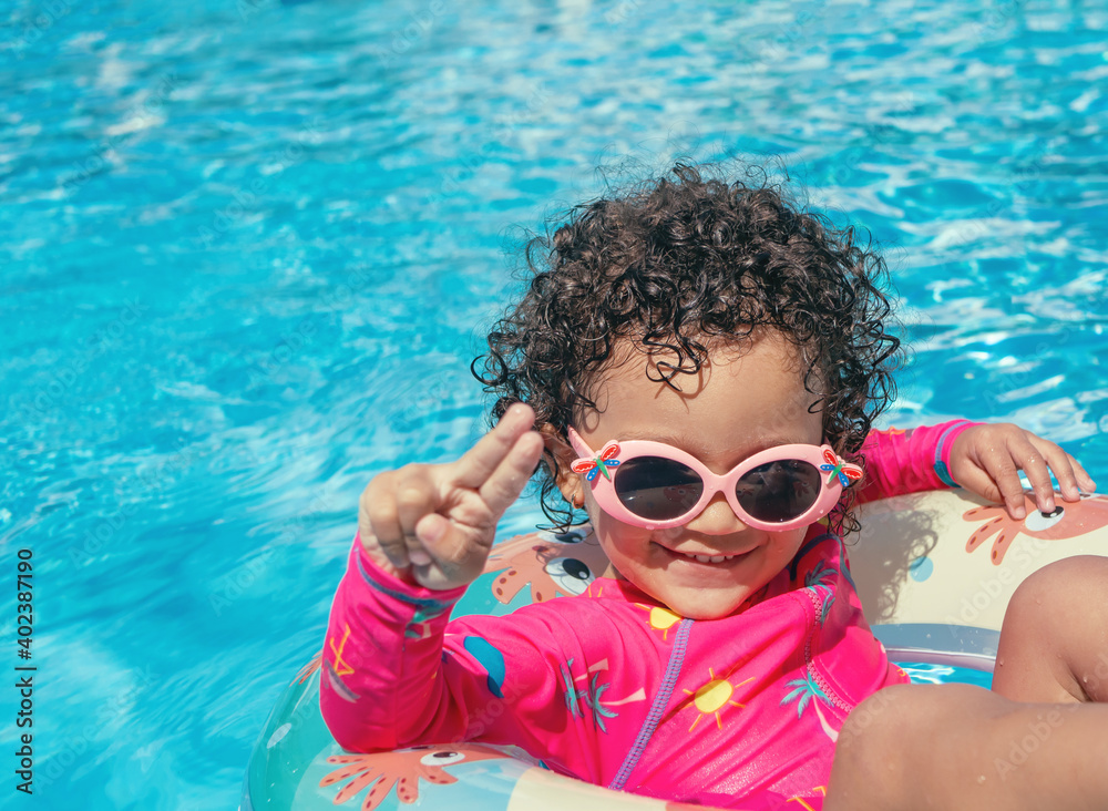Little girl with pink glasses, curly hair and fuchsia swimsuit, enjoys the blue water pool with her round float on a sunny day
