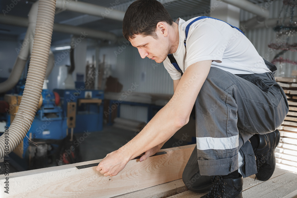 A young man works in a carpentry shop. An employee of Caucasian appearance in overalls measures wood with a ruler A young man works in a carpentry shop. An employee of Caucasian appearance in overalls measures wood with a ruler