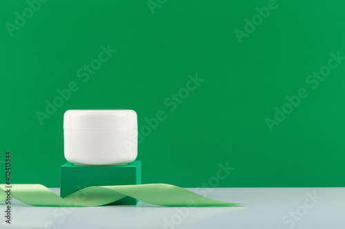 White jar with cream, mask or balm on green podium with ribbon on white glossy table against green background with copy space. Concept of organic cosmetics or daily skincare