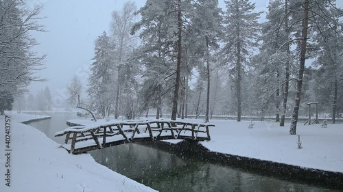 River flowing through stone canal with a wooden bridge in fairy tale winter landscape. Snowstorm in campground Sobec, Slovenia. Amazing view of snowflakes falling. Static shot, real time photo