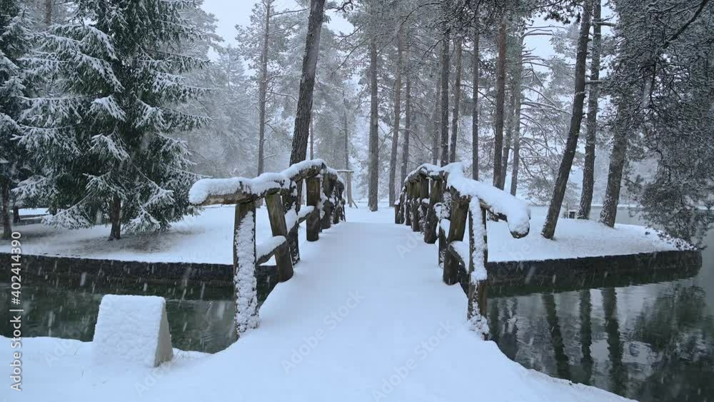 River flowing through stone canal with a wooden bridge in fairy tale winter landscape. Snowstorm in campground Sobec, Slovenia. Amazing view of snowflakes falling. Static shot, real time, wide angle River flowing through stone canal with a wooden bridge in fairy tale winter landscape. Snowstorm in campground Sobec, Slovenia. Amazing view of snowflakes falling. Static shot, real time, wide angle