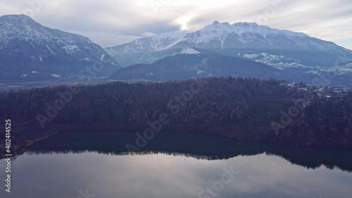 Drone aerial video footage above the Levico lake, facing the mountain Massiccio della vigolana, with sun setting in between the mountains behind the clouds. Flying above the water photo