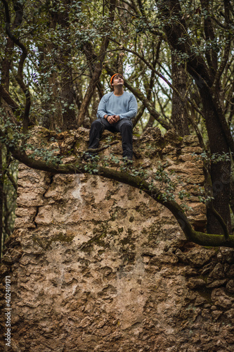 Young man hiking sitting on top of a wall of some ruins in dark mossy forest looking at the sky, vertical photo, man wears orange hat and teal sweater