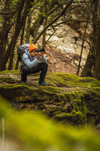 Young man with photo camera in the middle of the forest taking photos of the green and mossy landscape, man wears orange hat and teal sweater, green and brown Mediterranean landscape