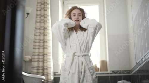 Portrait of positive redhead woman in bathrobe stretching in the morning in bathroom. Cheerful happy Caucasian lady waking up at home before shower. Weekend leisure concept.