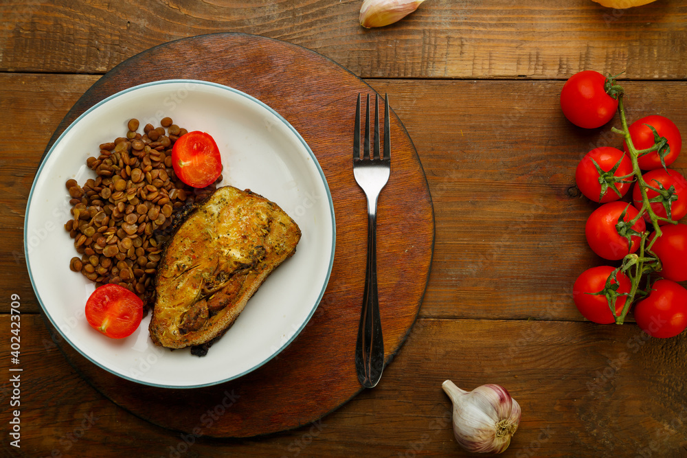 A plate of fish with lentils and tomatoes on a round stand with a fork near cherry tomatoes and lemon.