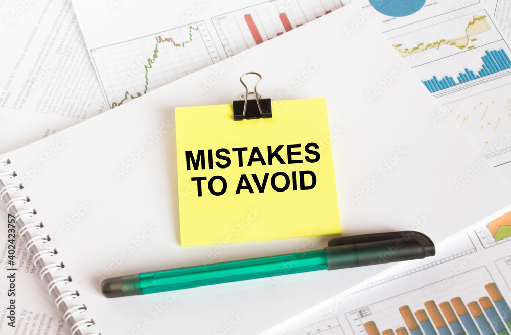 A yellow sticker with text Mistake To Avoid is in a Notepad with a green pen financial charts and documents A yellow sticker with text Mistake To Avoid is in a Notepad with a green pen financial charts and documents
