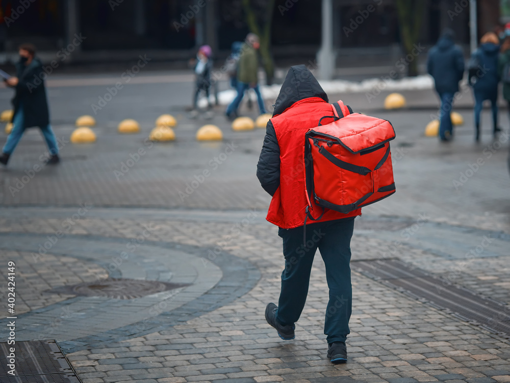 Obraz premium Delivery boy delivering online food orders to costumers, walking with red backpack, grocery delivery. Courier carries bag with fresh food. Delivering food at doorstep in holidays from restaurant