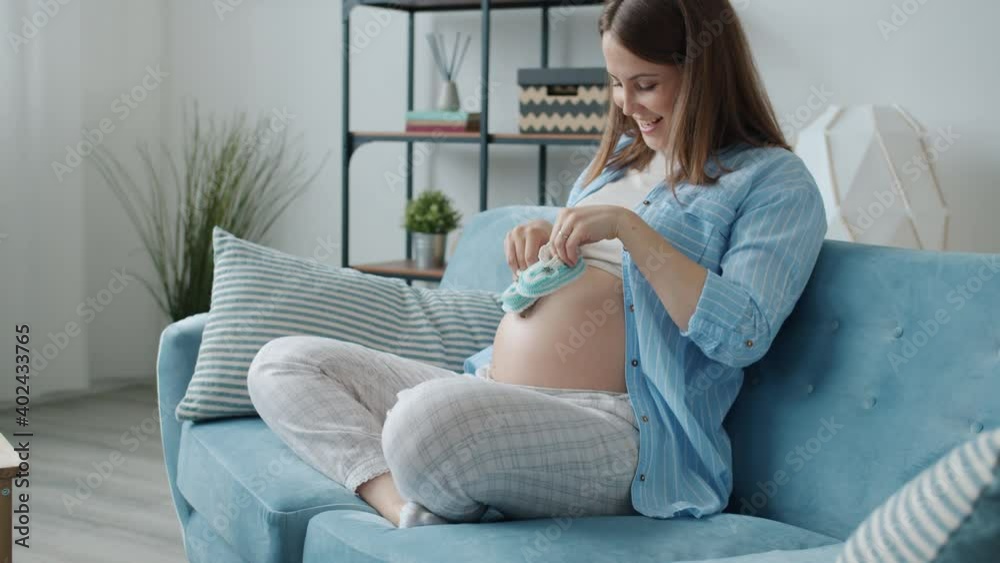 Cute pregnant woman is playing with blue baby booties walking on tummy having fun sitting on sofa alone at home. Pregnancy and happiness concept. Cute pregnant woman is playing with blue baby booties walking on tummy having fun sitting on sofa alone at home. Pregnancy and happiness concept.
