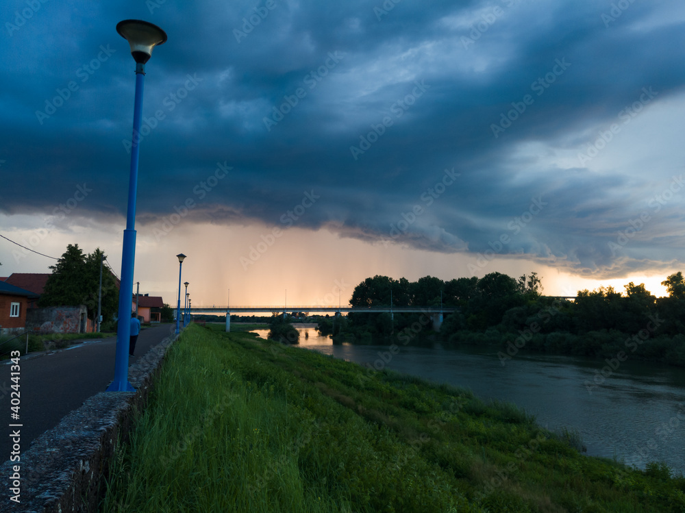 Storm dark cloud with heavy rain or summer shower, severe weather and sun glow behind rain. Landscape with Sava river and promenade in Bosanski Brod, Bosnia and Herzegovina during evening.