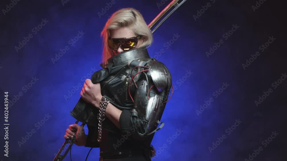Futuristic cyber woman weared with eyewear poses in dark blue background holding a sword on her shoulder looking at camera. Feminine authority and fantasy portrait of a futuristic warrior. Futuristic cyber woman weared with eyewear poses in dark blue background holding a sword on her shoulder looking at camera. Feminine authority and fantasy portrait of a futuristic warrior.