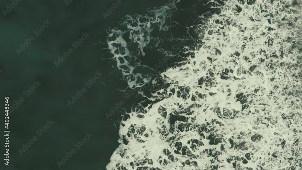 Aerial top down footage of large waves crashing in the shore; leaving a blanket of white foam behind
