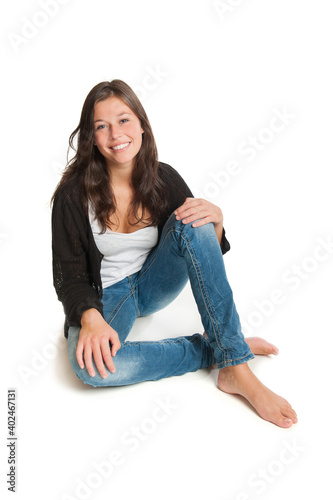 Laughing young woman wearing blue jeans, white top and a black wool jacket, she is sitting isolated in front of white studio background