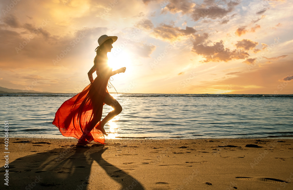 Young woman wearing long red dress and straw hat running on sand beach at sea shore enjoying view of rising sun in early summer morning. - obrazy, fototapety, plakaty 