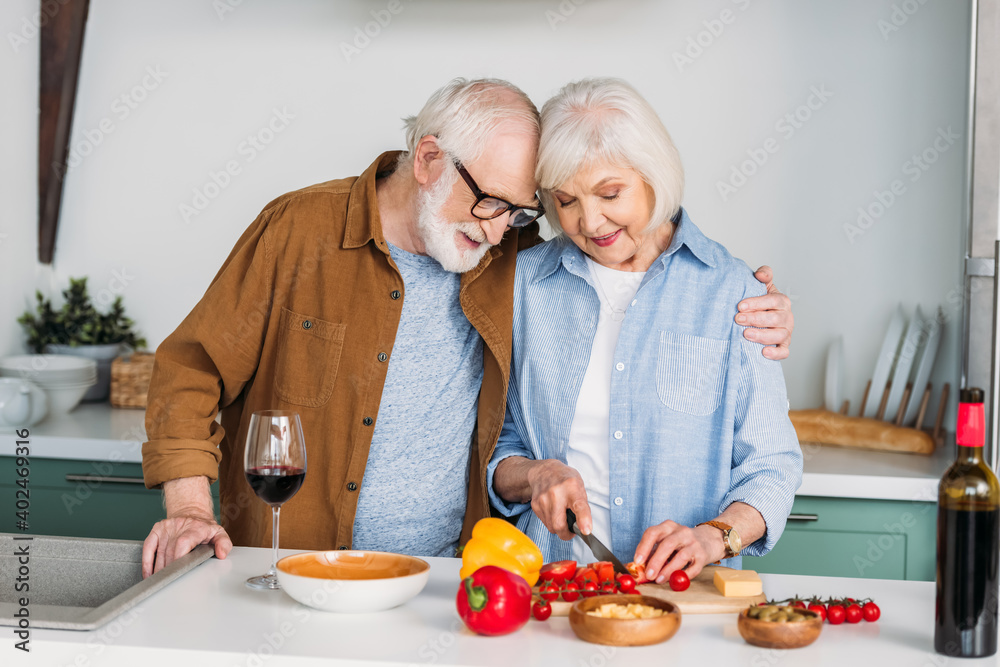 smiling senior husband hugging wife cooking dinner on table with wine, cheese and vegetables on blurred background smiling senior husband hugging wife cooking dinner on table with wine, cheese and vegetables on blurred background