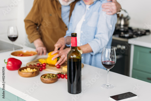 cropped view of elderly couple near table with bottle of wine and vegetarian food on blurred background