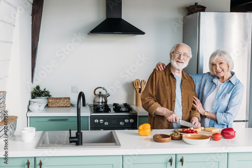 smiling senior wife looking at camera while hugging husband cooking dinner in kitchen