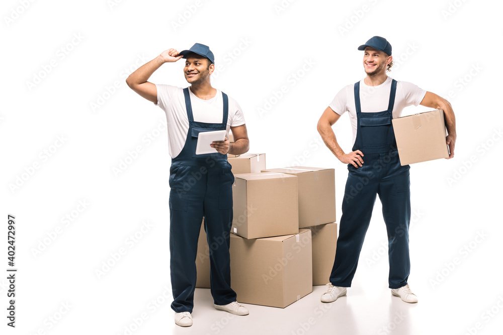 full length of cheerful indian worker holding digital tablet, adjusting cap and standing with mover near boxes on white