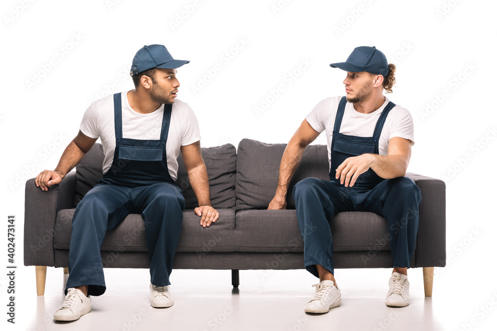 full length of shocked multicultural movers looking at each other while sitting on sofa on white