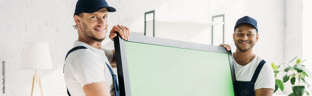 happy multicultural movers in uniform carrying plasma tv with green screen in apartment, banner happy multicultural movers in uniform carrying plasma tv with green screen in apartment, banner