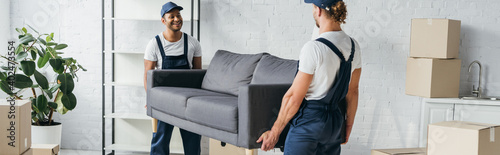 Fotografía smiling multiethnic movers in uniform carrying couch in apartment, banner