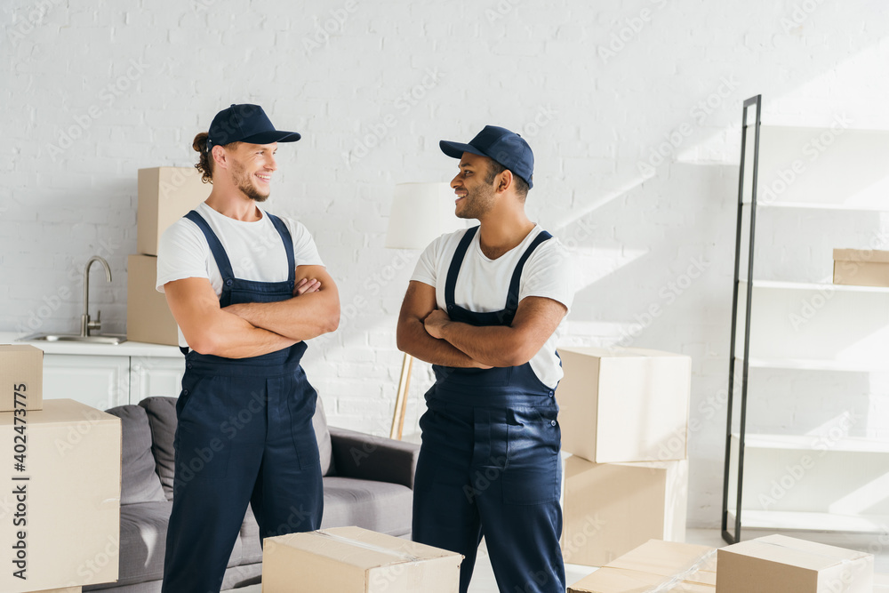 cheerful movers in caps standing with crossed arms while looking at each other near boxes in room cheerful movers in caps standing with crossed arms while looking at each other near boxes in room