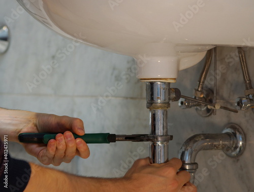 A close up of a hand holding a plumbing spanner screwing a fitting on wash basin waste pipe. 