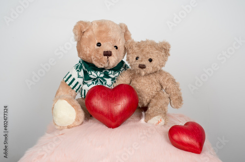 Two teddy bears sitting together on a pink cushion and holding a big wooden red heart as a couple in love with a smaller red heart placed on the right part of the cushion and a light grey background