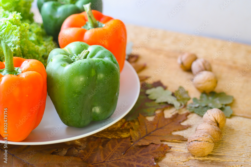 Bell peppers and lettuce leaves are on a plate. Around on a wooden background are yellowed oak leaves and walnuts in shell