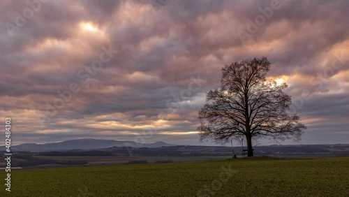 Timelapse of a bench and a lone tree on top of a hill overlooking dark colored clouds moving in the sky and overlooking a mountainous landscape.