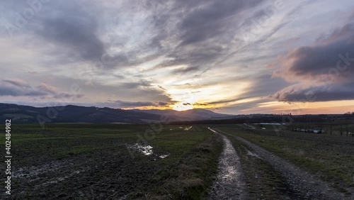 Timelapse of a dirt road and reflections on a road from sunset which sets behind a hilly landscape in a sky of colorful fast moving clouds.