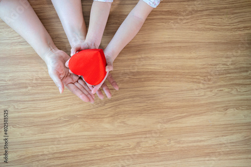 A red heart gift box in the hands of the child and mother on a white wooden background The concept of love