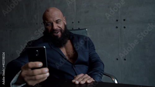 a muscular, bald, bearded man in a black shirt sits at a Desk and uses a mobile phone, talking on a video link and smiling.