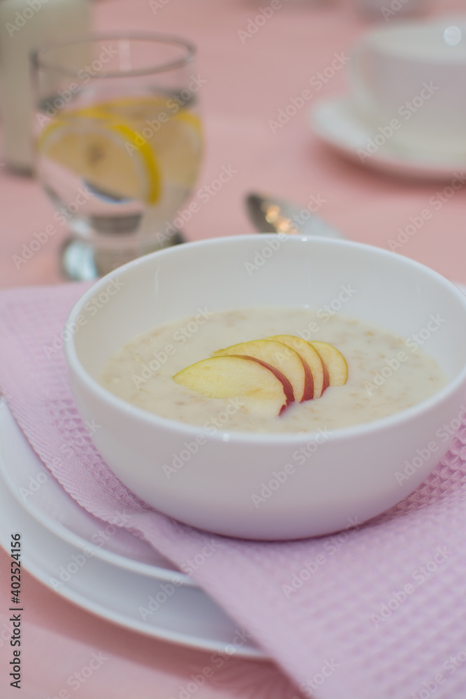 Oatmeal with apple for breakfast. Healthy eating. A useful breakfast. Porridge in a white ceramic cup on a pink tablecloth.