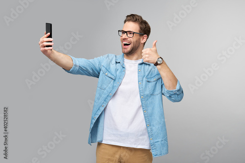 Smiling young man wearing jeans shirt taking selfie photo on smartphone or making video call standing over grey studio background