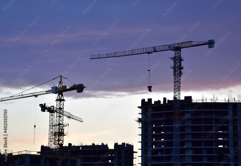 Silhouettes of tower cranes on awesome sunset background. Preparing to pour a bucket of concrete into formwork. Construction a multi-storey residential building. Small sharpness, possible granularity Silhouettes of tower cranes on awesome sunset background. Preparing to pour a bucket of concrete into formwork. Construction a multi-storey residential building. Small sharpness, possible granularity