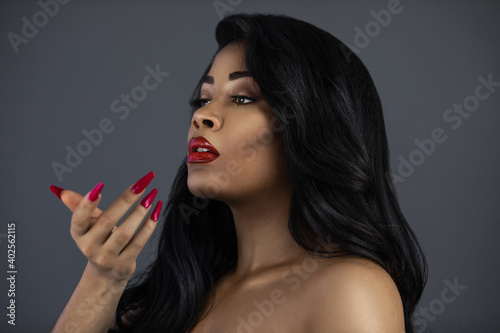 Fotografie, Obraz A portrait of a sensual young Latina with long black curly hair, beautiful makeup, red lips and nails posing by herself in a studio with a grey background