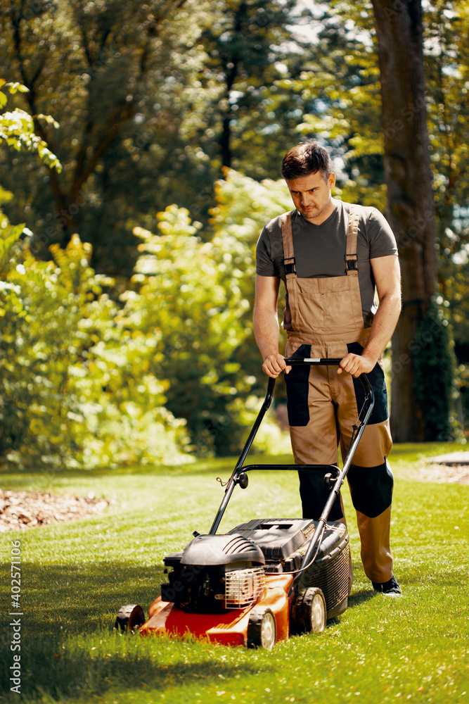 Young handsome gardener waring brown overall and mowing the grass Young handsome gardener waring brown overall and mowing the grass