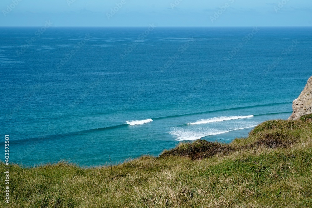 A small blue wave goes unridden behind the grassy cliff, Cornwall