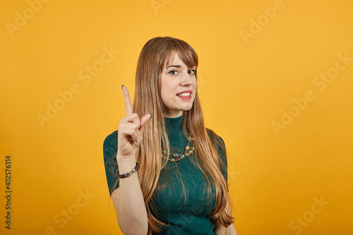 Hand points index finger up, pointing forefinger surprise gift option, great idea. Young attractive woman, dressed green shirt blonde hair, yellow background