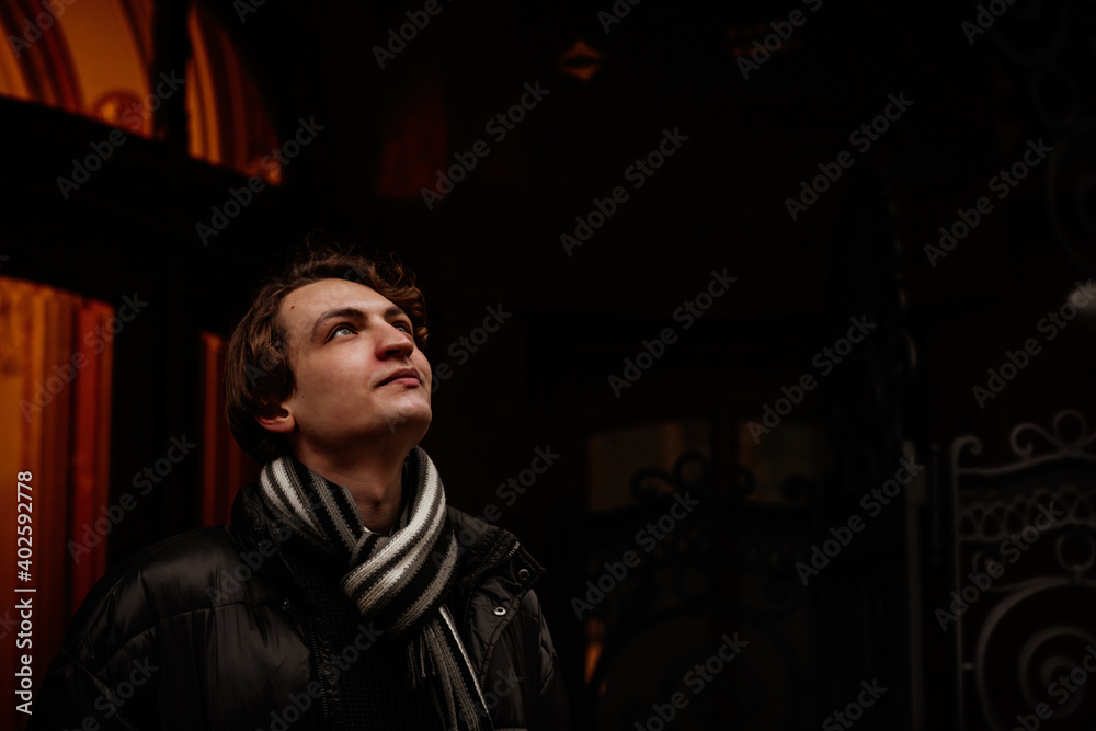 Obraz premium Handsome stylish young man wearing winter clothes in street. Portrait of beautiful guy with modern hairstyle with jacket and scarf. Vintage European city, lights on background. Soft focus.