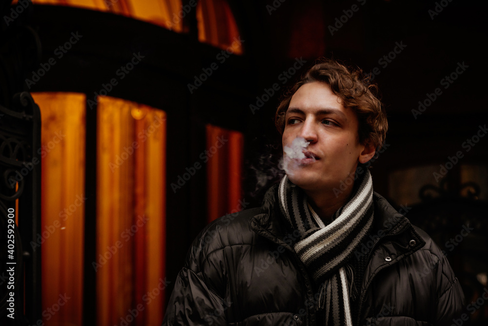 Handsome stylish young hipster man smoking cigarette on the street in winter, looking away. Portrait of beautiful guy dressed casually. Vintage European city, lights on background. Soft focus. Handsome stylish young hipster man smoking cigarette on the street in winter, looking away. Portrait of beautiful guy dressed casually. Vintage European city, lights on background. Soft focus.