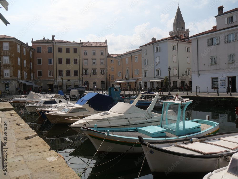 Mandracchio in Muggia : it is a small harbor inside the village and surrounded by ancient walls and colorful buildings. There are colored boats moored and reflected in the water.
