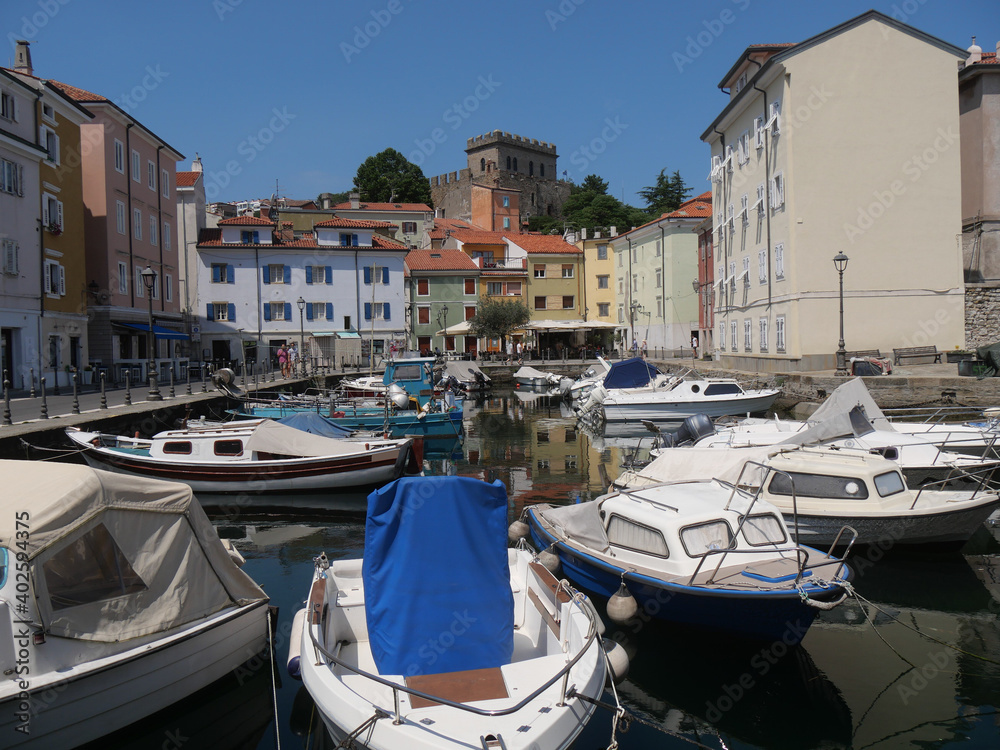 Mandracchio in Muggia : it is a small harbor inside the village and surrounded by ancient walls and colorful buildings. There are colored boats moored and reflected in the water.