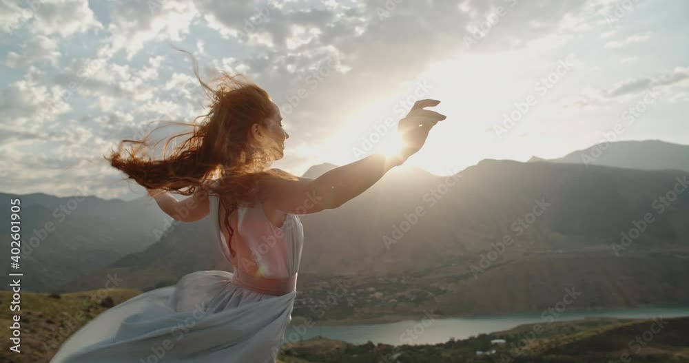 Woman in white dress standing on top of a mountain with raised hands while wind is blowing her dress and red hair - freedom, nature concept 4k footage