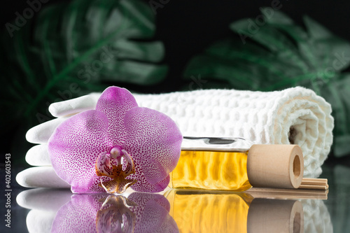 Fototapeta Naklejka Na Ścianę i Meble -  Lilac orchid flower, clear bottle of yellow oil, wooden sticks and towel with white stones and monstera on black surface