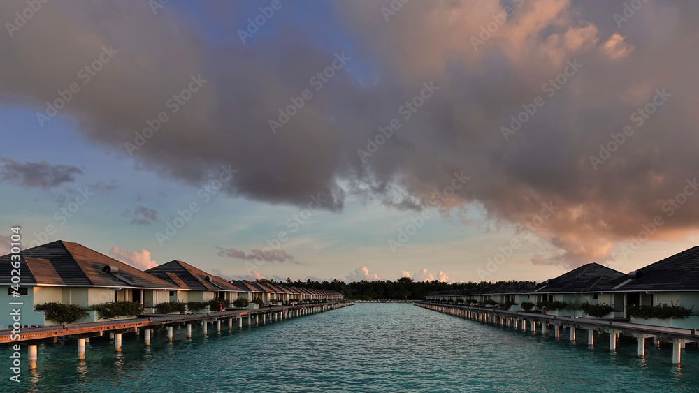 Dawn in the Maldives. There are pink cumulus clouds in the sky. The turquoise ocean is calm. Two rows of villas stretch over the water to the coast. No people