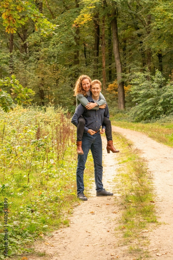 Attractive young couple, man carries woman on his back in the woods. They stand still on a sandy forest path Attractive young couple, man carries woman on his back in the woods. They stand still on a sandy forest path