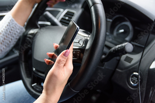 Young woman in jeans and blue sweater is driving the car with one hand, using her smartphone by another hand. Extremely unsafety and risky driving. © Дмитрий Березнев